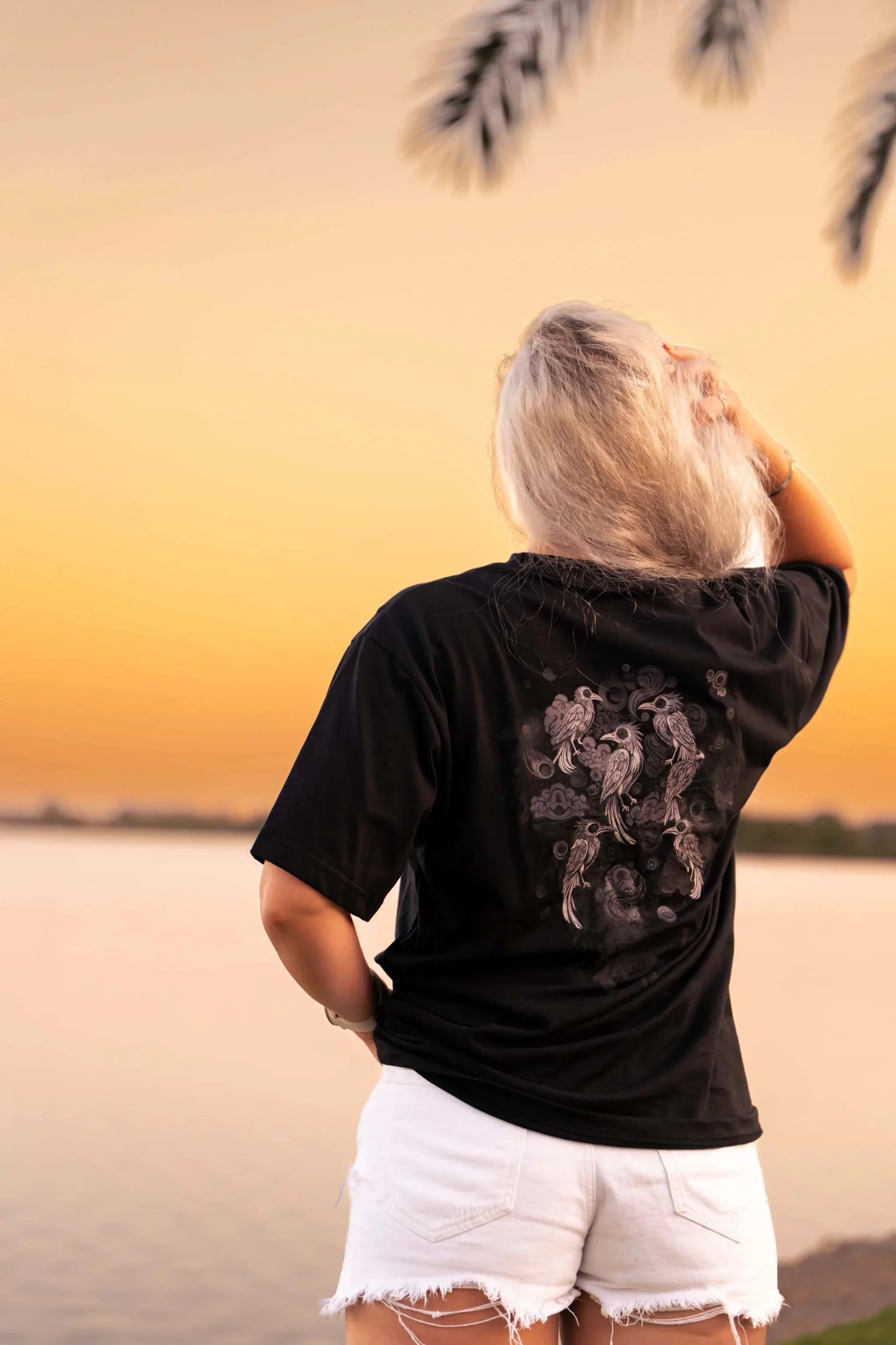Persona de espaldas usando camiseta negra Cuervos Nocturnos con diseño gótico al atardecer junto al agua