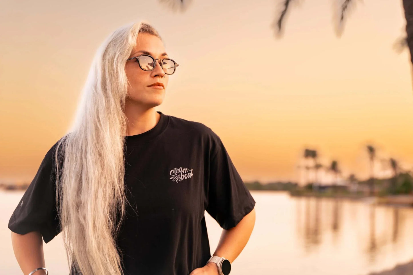 Mujer con gafas y cabello largo rubio viste camiseta negra Cuervos Nocturnos estilo gótico al atardecer.