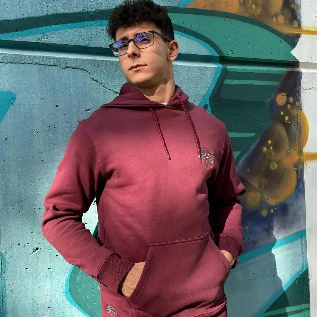 Hombre con sudadera Rose unisex burdeos minimalista con capucha y bolsillo canguro frente a muro grafiteado.