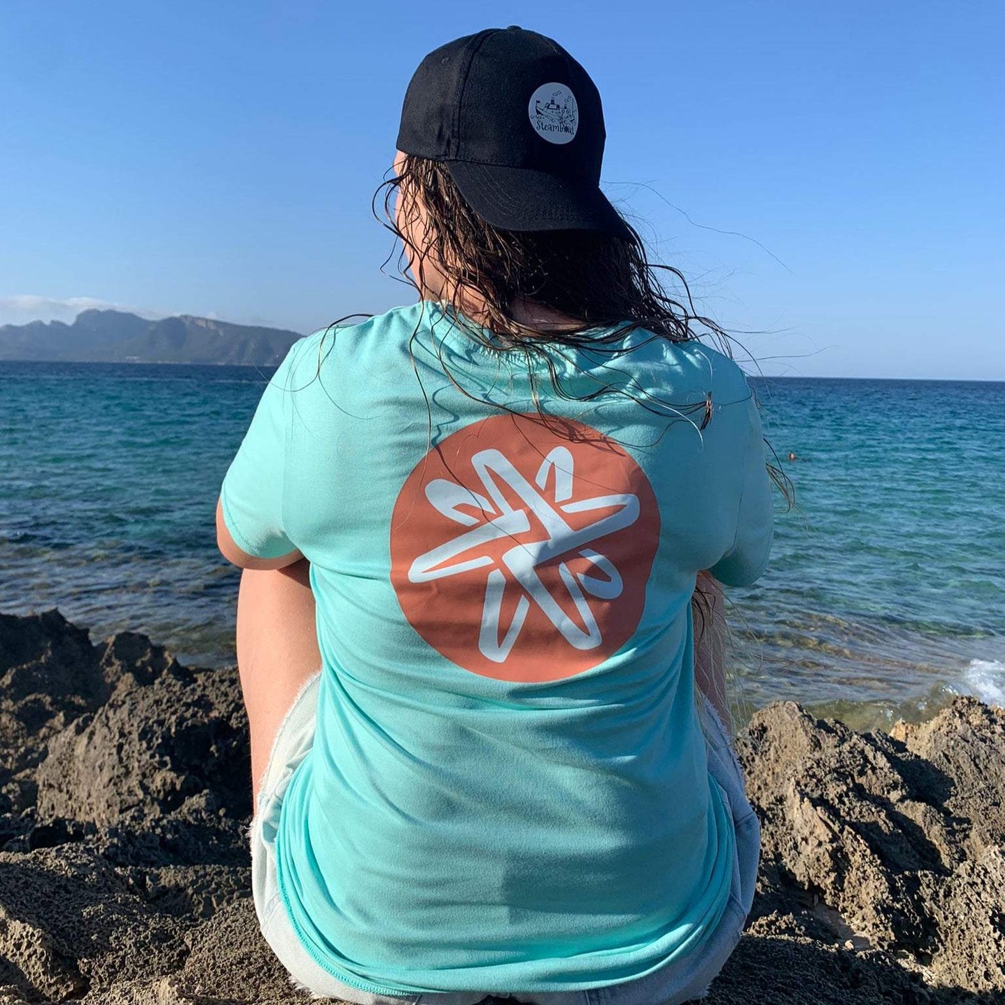 Mujer de espaldas con camiseta básica verde menta unisex y gorra negra junto al mar y rocas