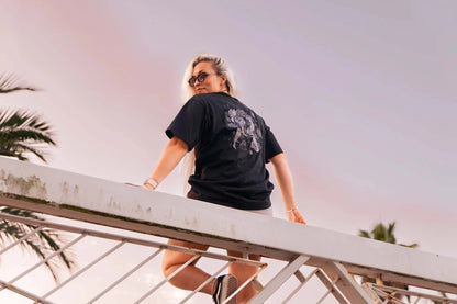 Persona con camiseta Cuervos Nocturnos negra con diseño gótico y estampado oscuro vista desde atrás en exterior al atardecer